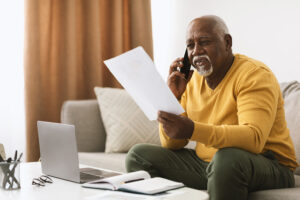 Senior Black Businessman Talking On Mobile Phone Holding Papers Working Sitting At Laptop Indoors. Mature African American Man Speaking By Cellphone In Living Room At Home. Communication Concept